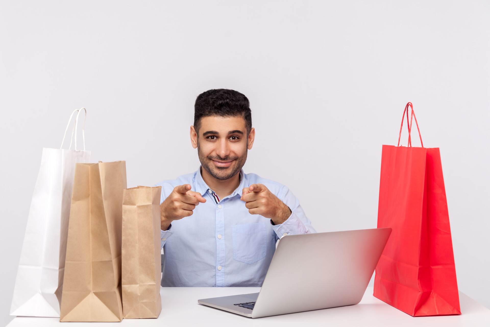 Homme beau gai s’asseyant au bureau avec l’ordinateur portatif, entouré des paquets, pointant au sourire de caméra, magasin en ligne